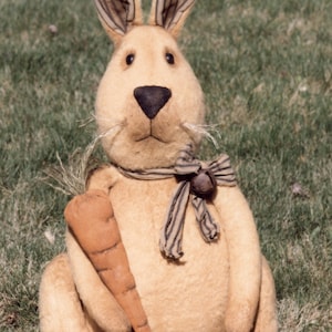May include: A plush brown bunny rabbit with black and white striped ears, holding a carrot in its paws. The bunny is wearing a black and white striped ribbon with a bell around its neck. The bunny is sitting on a bed of green grass.