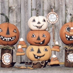 Può includere: Tre decorazioni di zucca dipinte con accenti di mais dolce. Le zucche sono impilate l'una sull'altra e hanno volti dipinti. Le zucche sono su una superficie di legno. Il testo "got candy?" è su un cartello attaccato a una delle zucche.