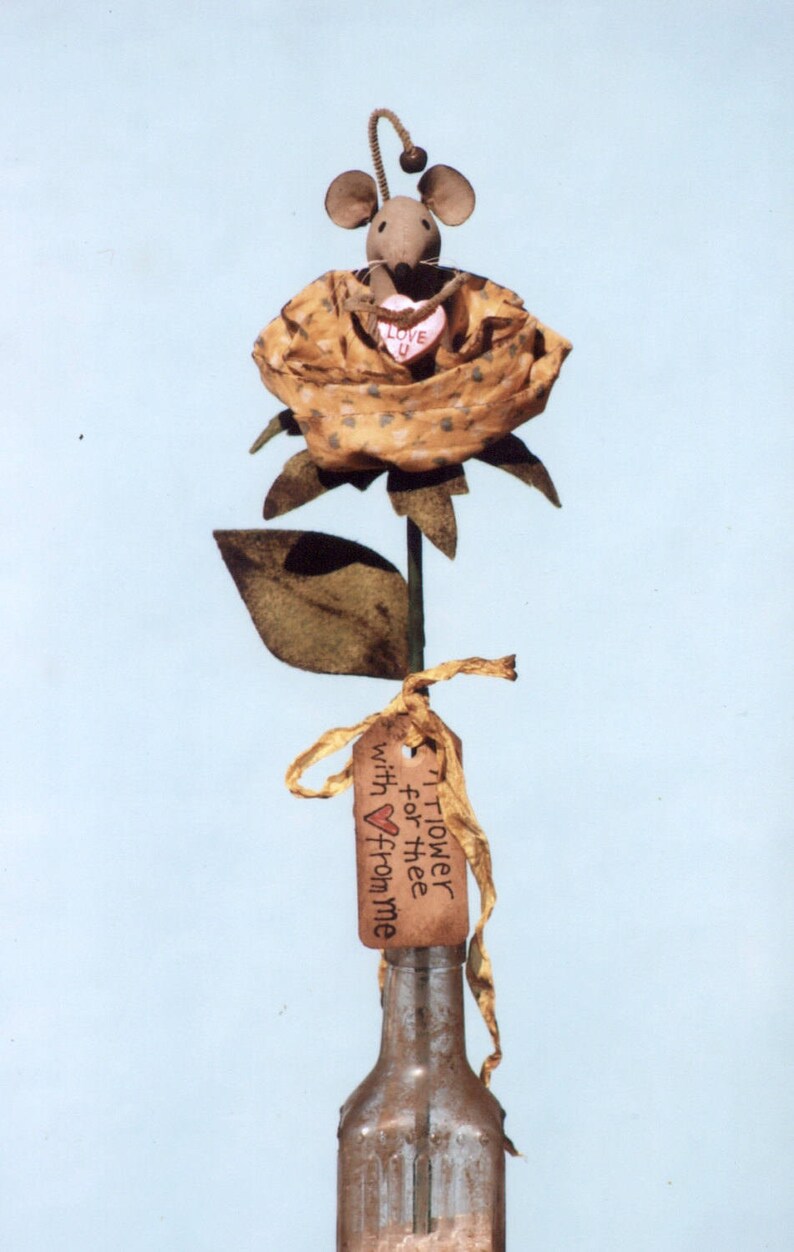 May include: A brown mouse with a pink heart that says "Love U" sits on top of a brown fabric rose with a brown stem and leaves. A brown tag with the text "A Flower for thee with &hearts; from me" is tied to the stem.