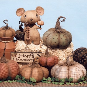 May include: A collection of handmade patchwork pumpkins with a brown mouse figurine holding a miniature pumpkin. The pumpkins are decorated with different patterns and colors, including brown, green, and white. A sign reads "Happy Harvest".