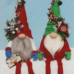 Könnte beinhalten: Zwei Weihnachtsgnome, einer mit roter Mütze und der andere mit grüner Mütze, sitzen auf einer weißen Oberfläche. Beide Gnomen haben weiße Bärte und tragen rote Hosen. Der Gnom mit der roten Mütze hält ein Schild mit der Aufschrift "Birds Needed".