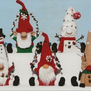 Pode incluir: Cinco gnomos de Natal feitos à mão, incluindo um boneco de neve, uma rena e três gnomos com chapéus vermelhos, verdes e brancos. Os gnomos usam guirlandas e têm barbas brancas. Eles estão sentados em uma superfície nevada.