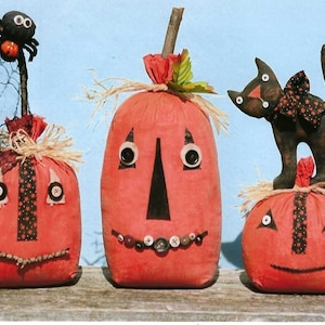 Puede incluir: Tres calabazas de tela hechas a mano con caras blancas y negras. La calabaza de la izquierda tiene una ara&ntilde;a en la parte superior. La calabaza del medio tiene una fila de botones para los dientes. La calabaza de la derecha tiene un gato negro de pie en la parte superior.
