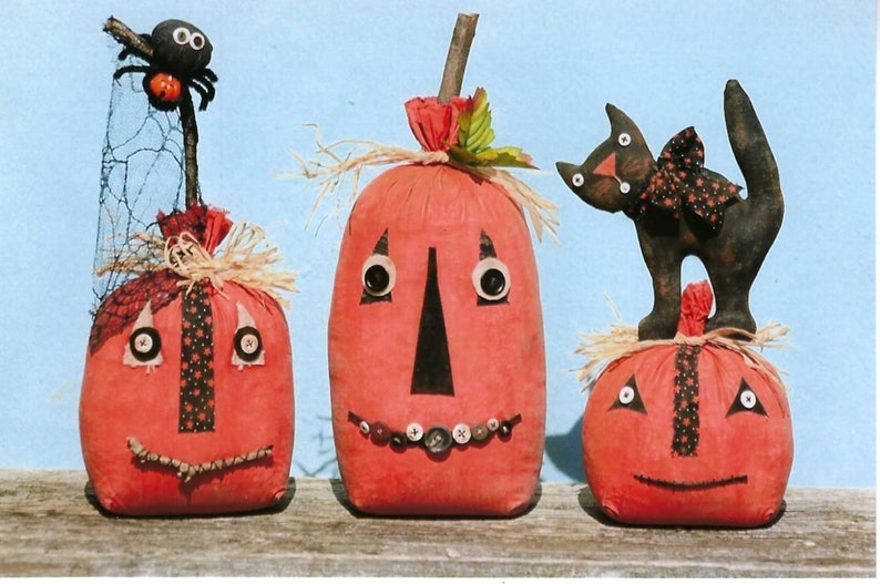 Puede incluir: Tres calabazas de tela hechas a mano con caras blancas y negras. La calabaza de la izquierda tiene una ara&ntilde;a en la parte superior. La calabaza del medio tiene una fila de botones para los dientes. La calabaza de la derecha tiene un gato negro de pie en la parte superior.
