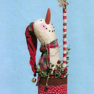 May include: A snowman figurine with a red hat, plaid scarf, and a candy cane. The snowman is standing in a brown pot with a red and white candy cane pattern. The snowman is decorated with green sprigs and red berries.