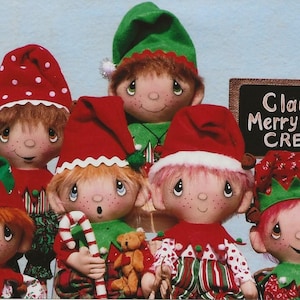 May include: A group of six handmade elf dolls dressed in festive Christmas attire. The elves are wearing red and green hats, shirts, and pants with white polka dots. The dolls have painted faces with rosy cheeks and big eyes. A chalkboard sign reads "Claus' Merry Little Crew".