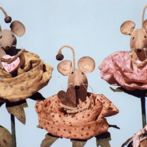 May include: Three handmade fabric mouse figurines, each holding a heart-shaped decoration. The mice are dressed in fabric roses, one yellow, one pink, and one orange. The hearts have the words "Love You" and "Love" printed on them.