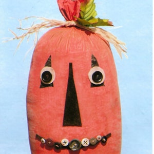 Puede incluir: Una bolsa de tela naranja en forma de calabaza hecha a mano con un tallo de palo y una cara de fieltro negro. La cara tiene dos ojos de fieltro negro con botones blancos, una nariz de fieltro negro y una fila de botones negros para la boca.
