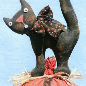 Puede incluir: Un gato negro con un lazo rojo y blanco de lunares est&aacute; de pie sobre una calabaza roja y negra con paja alrededor de la base.