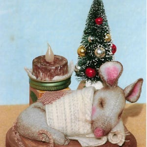 May include: A handcrafted Christmas scene featuring a sleeping mouse figurine on a wooden base. A small decorated Christmas tree and a candle are also present. The mouse is gray with pink ears and nose, and is covered with a white blanket.