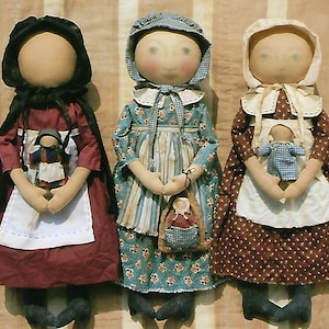 May include: Three handmade fabric dolls wearing bonnets and dresses. The dolls are holding smaller dolls. The dolls are standing on a striped fabric background.