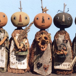 May include: Five handmade pumpkin-shaped dolls with different facial features and clothing. Each doll is holding a different autumnal item, such as sunflowers, wheat, and a basket of leaves. The dolls are wearing brown, beige, and white clothing. The text on the dolls' clothing reads "Gathering Time", "A Time To Gather", and "Gather".