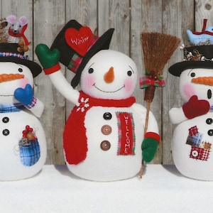 May include: Three handmade snowman figurines with different hats and accessories. The snowman on the left has a blue plaid scarf, a blue and white hat with a mouse on top, and a red berry sprig. The middle snowman has a black top hat with a red heart that says "Winter" on it, a red scarf, and a broom. The snowman on the right has a black top hat with a bird's nest on top, a blue plaid scarf, and a red mitten.