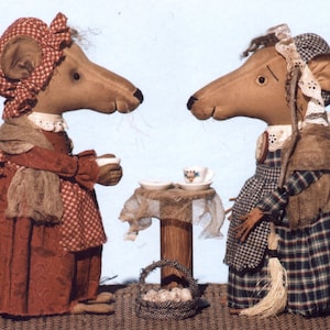 May include: Two brown fabric mouse dolls wearing dresses and bonnets are sitting at a small table with teacups and a basket of eggs. The mouse on the left is wearing a red and white checkered dress with a brown shawl. The mouse on the right is wearing a blue and white plaid dress with a brown shawl.