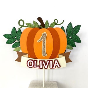 May include: Orange pumpkin cake topper with green leaves, a brown stem, and a glittery gold number 1. A white banner with the name "Olivia" in red letters is attached to the bottom of the pumpkin.