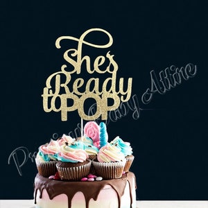 Puede incluir: Una tarta de revelación de género con un adorno dorado brillante que dice "She's Ready to POP". La tarta tiene una base azul, blanca y rosa, con glaseado de chocolate goteando por los lados. Cupcakes con glaseado rosa y azul coronan la tarta.
