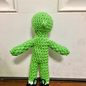 May include: A green crocheted doll with black and white shoes. The doll is standing on a brown surface.