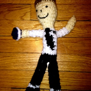 May include: A crocheted doll with a smiling face, wearing a white and black shirt and black pants. The doll is standing on a wooden surface.