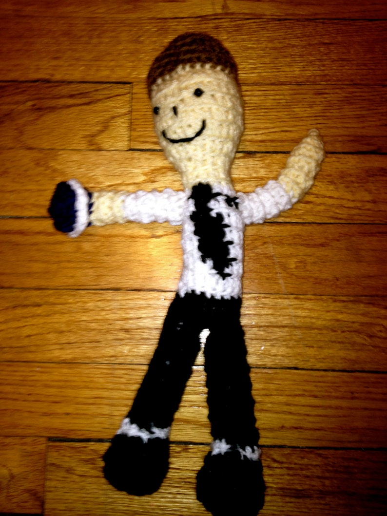 May include: A crocheted doll with a smiling face, wearing a white and black shirt and black pants. The doll is standing on a wooden surface.