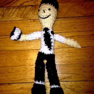 May include: A crocheted doll with black hair, a white shirt with a black tie, and black pants. The doll is smiling and has a black and white pattern on its shirt.