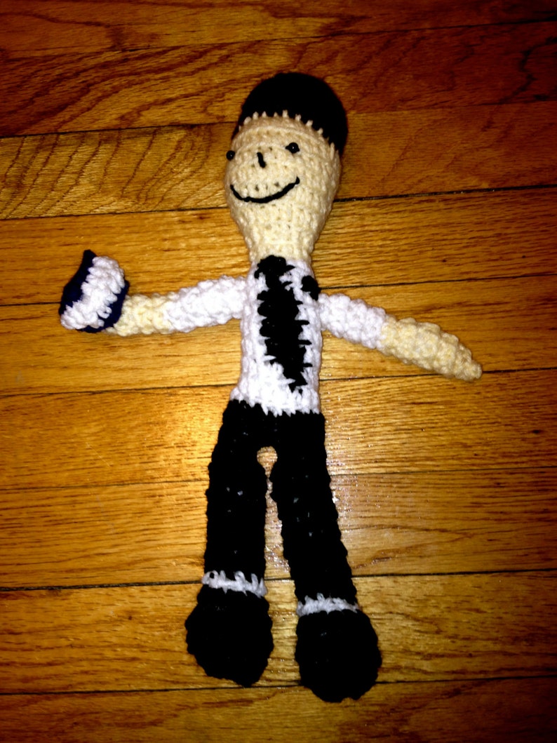 May include: A crocheted doll with black hair, a white shirt with a black tie, and black pants. The doll is smiling and has a black and white pattern on its shirt.