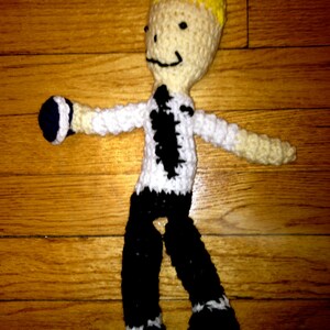 May include: A crocheted doll wearing a white shirt with a black tie, black pants, and black shoes. The doll has yellow hair and a smiling face.