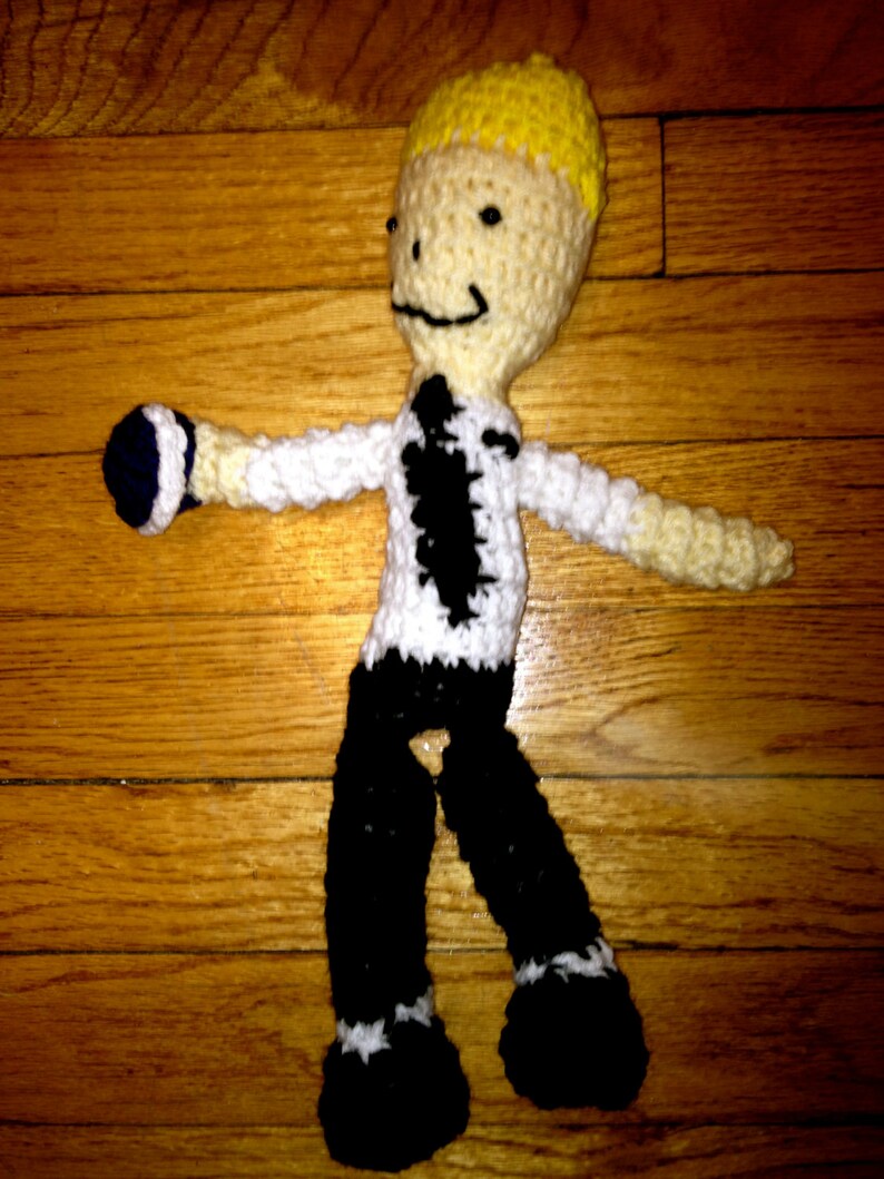 May include: A crocheted doll wearing a white shirt with a black tie, black pants, and black shoes. The doll has yellow hair and a smiling face.