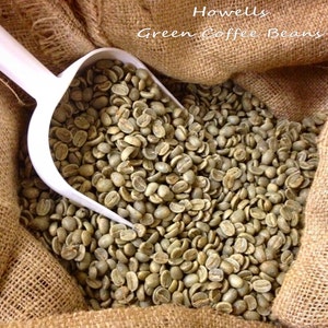 May include: A burlap sack filled with green coffee beans. A white scoop is partially filled with beans and is being lifted out of the sack. The text "Howells Green Coffee Beans" is printed on the sack.