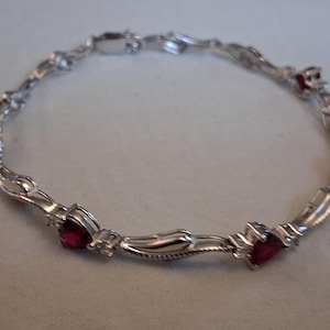 May include: A silver-toned metal link bracelet featuring alternating sections of polished metal and small, heart-shaped red gemstones. The bracelet is designed with a repeating pattern of curved metal links and clear accent stones set near each red heart.