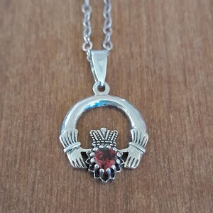 May include: Silver Celtic Claddagh necklace with a red gemstone heart. The hands are clasped together and the crown is on top of the heart.