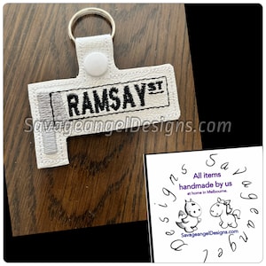Pode incluir: Chaveiro branco com um nome bordado em preto "RAMSAYST" em um fundo branco. O chaveiro tem um anel prateado e um botão branco.