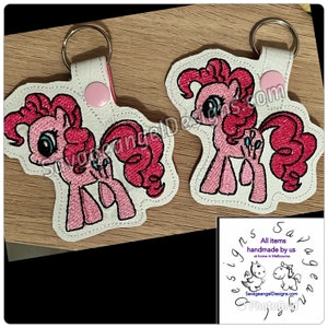 May include: Two pink and white embroidered keyrings featuring a cartoon pink pony with a blue eye. The keyrings are on a wooden surface. The text "SavageangelDesigns.com" is visible in the image.