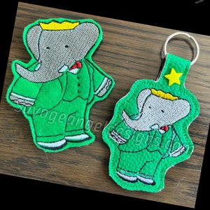Peut inclure: Un porte-clés en feutre vert avec un éléphant brodé gris portant un costume vert et une couronne jaune. L'éléphant a un nœud papillon rouge et des chaussures blanches. Le porte-clés a une étoile dorée et un anneau métallique.