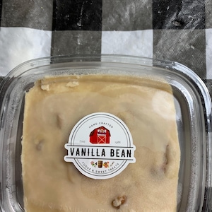 May include: A square clear plastic container holding a block of light tan vanilla bean fudge. A circular white label on the lid features a red barn illustration, the text "HAND CRAFTED", "ESTD 1986", "VANILLA BEAN", and "FUDGE & SWEET TREATS".