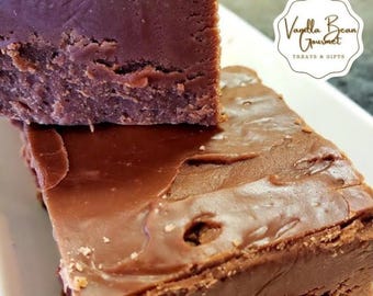 May include: Two stacked squares of chocolate fudge with a smooth, glossy top layer and a dense, textured base. A white logo sticker reads Vanilla Bean Gourmet TREATS & GIFTS, and the text WWW.VANILLABEANINC.COM is printed at the bottom.