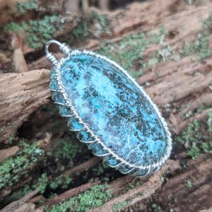 Op de afbeelding: Een turquoise blauwe stenen hanger met een zilveren draadwikkeling. De steen heeft een gevlekt patroon en is in een ovale vorm gezet.