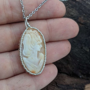 以下が含まれることがあります： 銀色のチェーンネックレスに、彫刻された女性の横顔が描かれたカメオペンダントが飾られています。カメオは銀色のワイヤーフレームにセットされています。