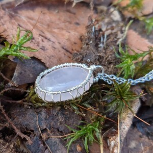 Op de afbeelding: Een zilveren ketting met een hanger van roze kwarts. De hanger is gewikkeld in zilverdraad en heeft een natuurlijke, ovale vorm.