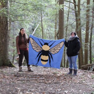 May include: Two people are holding up a blue crocheted blanket with a large yellow and black bumblebee design in a forest setting.
