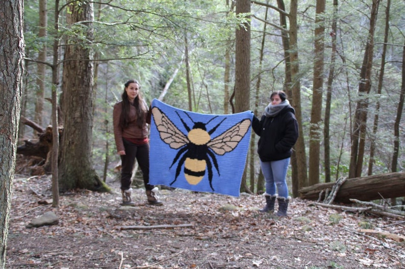 May include: Two people are holding up a blue crocheted blanket with a large yellow and black bumblebee design in a forest setting.