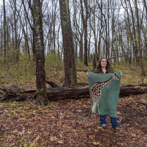 May include: A person is standing in a wooded area holding a crocheted blanket with a giraffe design. The blanket is green with brown and tan giraffe spots.