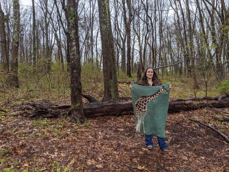 May include: A person is standing in a wooded area holding a crocheted blanket with a giraffe design. The blanket is green with brown and tan giraffe spots.
