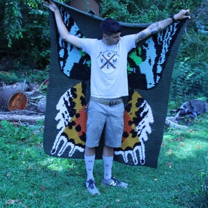 May include: A person holding a green, blue, and yellow crocheted blanket with a butterfly design. The blanket has the text "YCW" in the center.