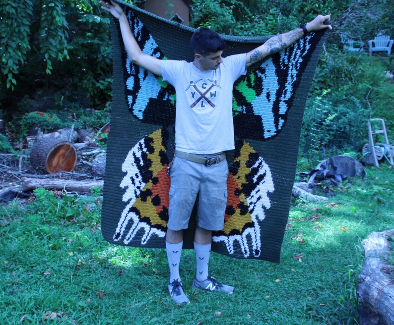 May include: A person holding a green, blue, and yellow crocheted blanket with a butterfly design. The blanket has the text "YCW" in the center.