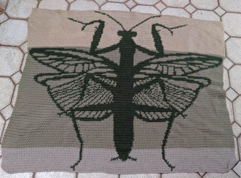 May include: A crocheted throw blanket featuring a green praying mantis with butterfly wings. The blanket is made with a beige and brown yarn.