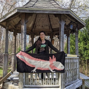 May include: A person holding a crocheted blanket featuring an axolotl, a type of salamander, in pink, white, and black colors. The blanket is displayed in front of a wooden gazebo.
