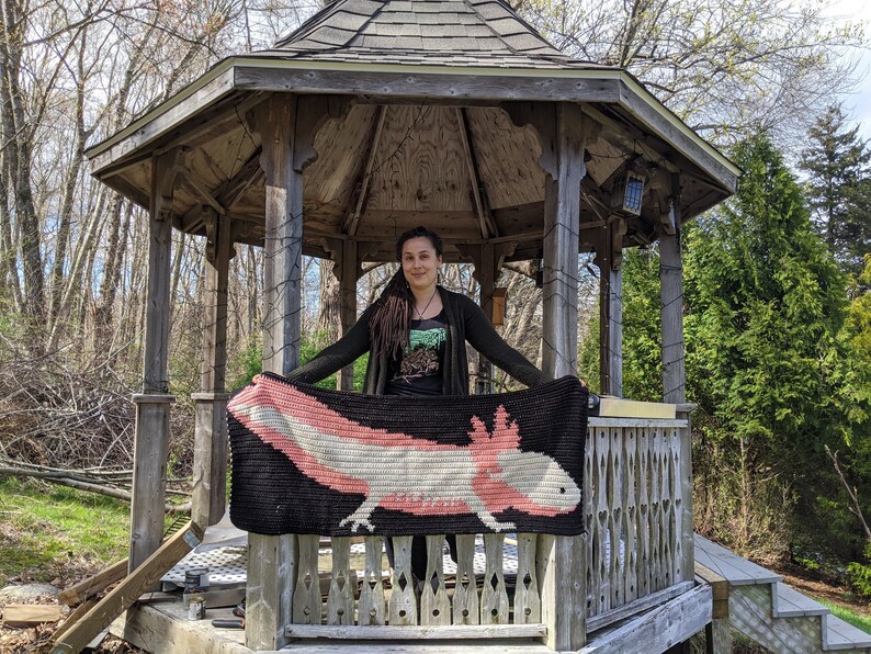 May include: A person holding a crocheted blanket featuring an axolotl, a type of salamander, in pink, white, and black colors. The blanket is displayed in front of a wooden gazebo.