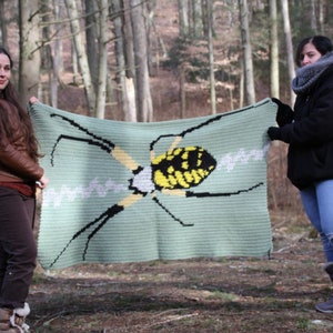 May include: Two people are holding up a crocheted blanket with a yellow and black spider design. The blanket is green and has a white web design.