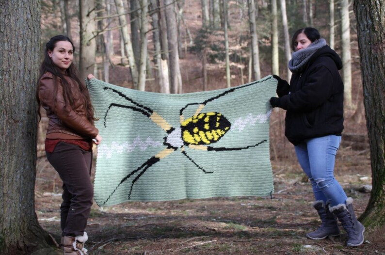 May include: Two people are holding up a crocheted blanket with a yellow and black spider design. The blanket is green and has a white web design.