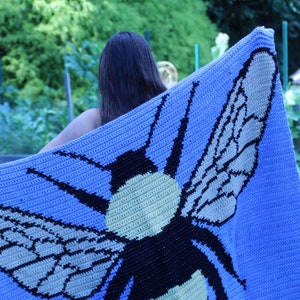 May include: A crocheted blanket featuring a large bee design. The bee is black, yellow, and white, with wings that are beige and black. The blanket is a light blue color.